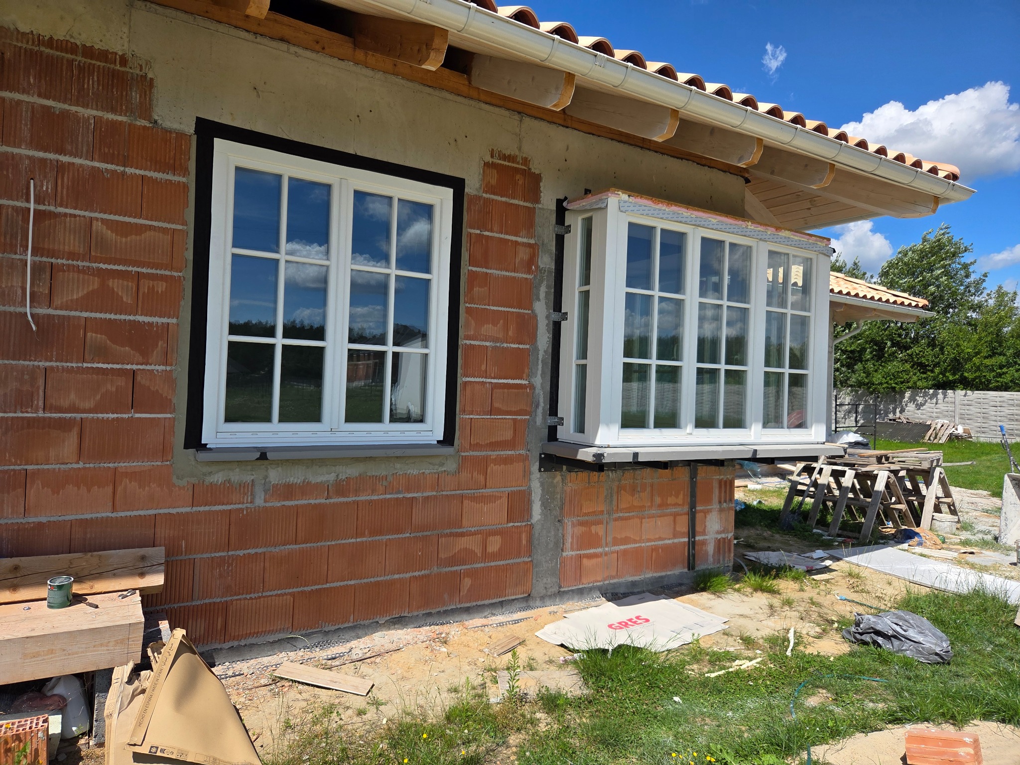 Dwa białe okna z szybami zamontowane w surowej ścianie budynku z czerwonej cegły. Jedno okno jest standardowe, drugie wystaje na zewnątrz.