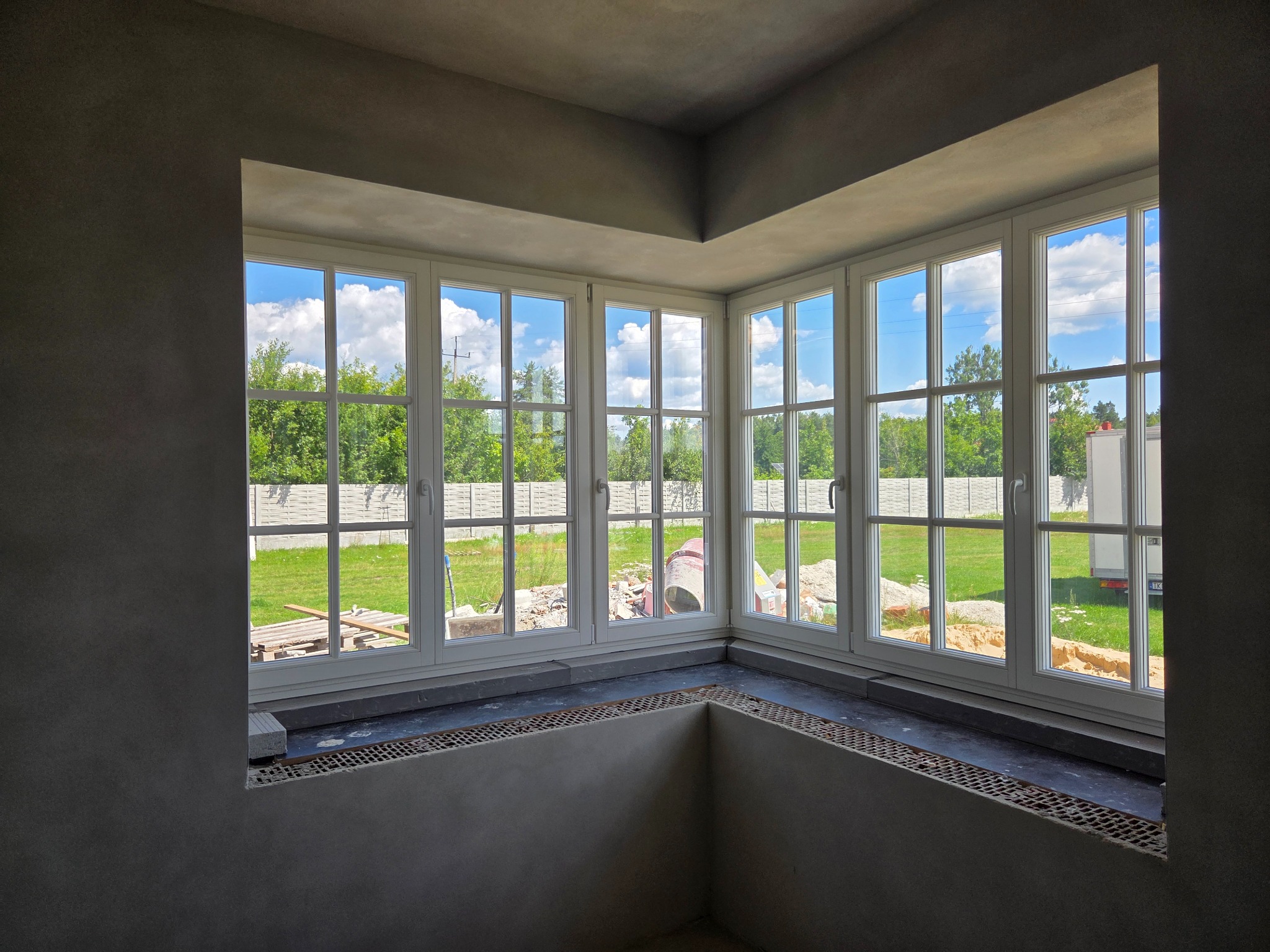 Wnętrze z surowymi ścianami i narożnym oknem z białymi ramami, przez które widać zielony ogród i błękitne niebo z chmurami. Widoczne przygotowania do wykończenia.