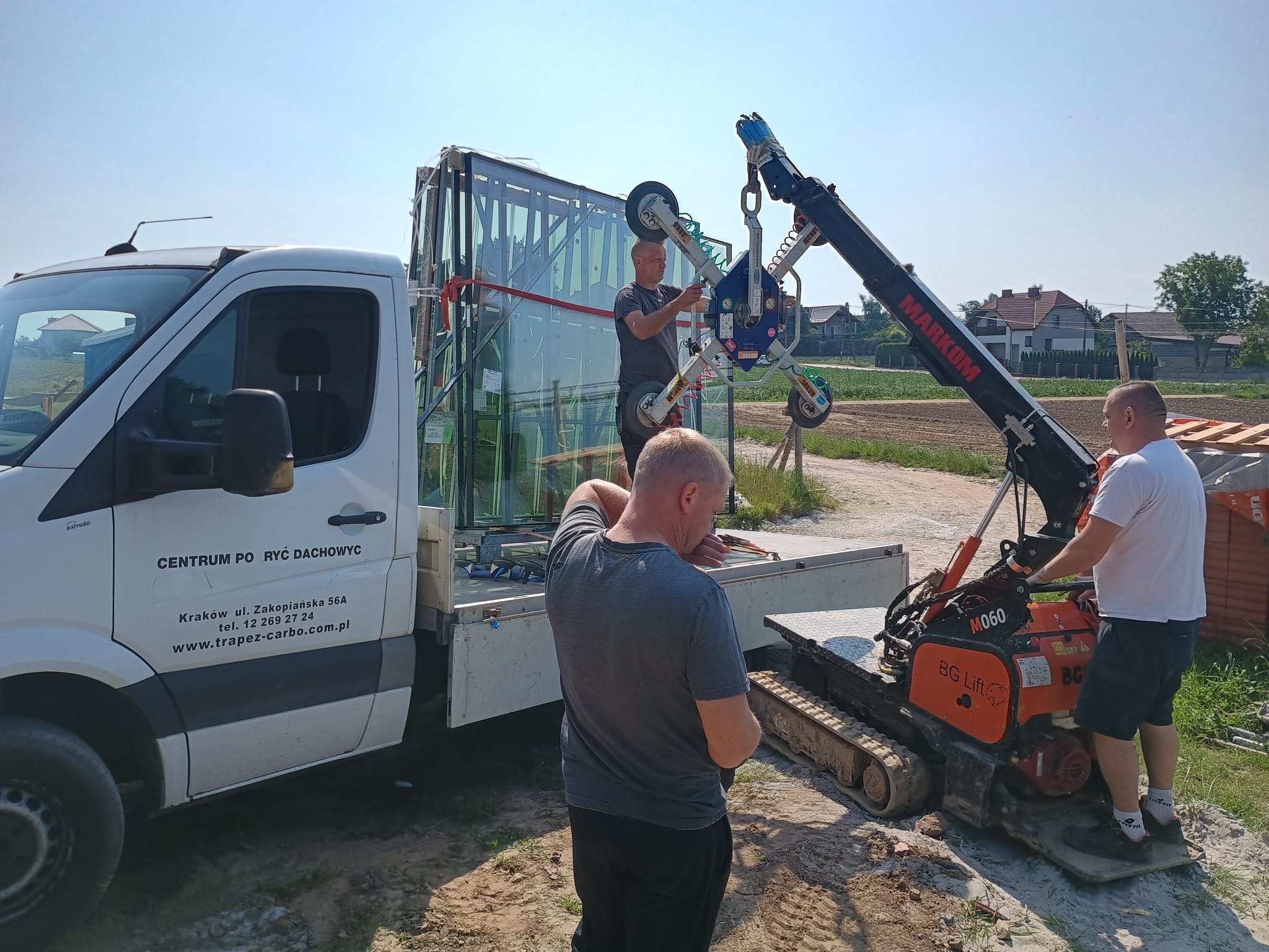 Montaż dużych okien z użyciem dźwigu przyssawkowego. Pracownicy w trakcie operacji na tle firmowego samochodu dostawczego i zabudowy jednorodzinnej.