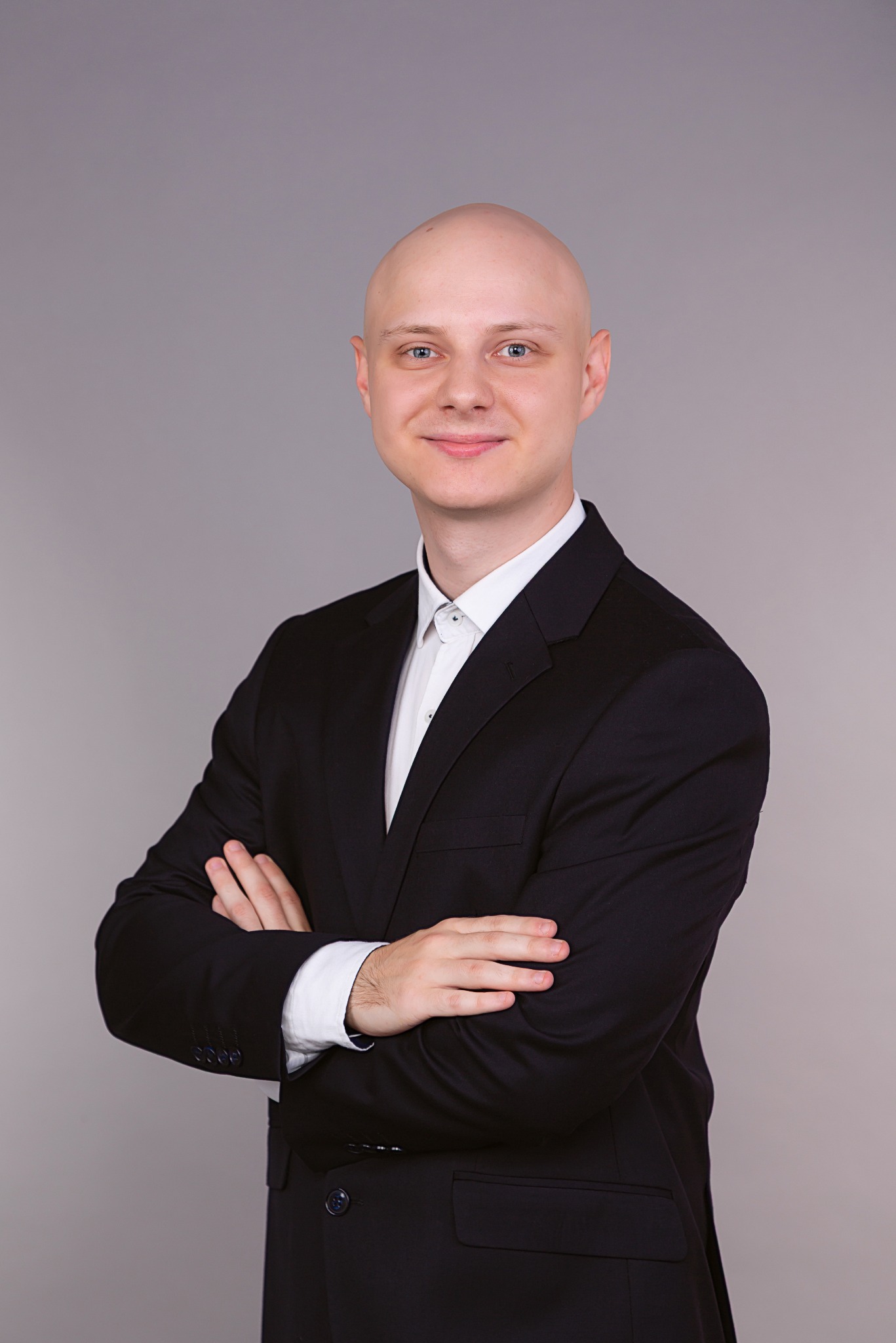 Portret uśmiechniętego mężczyzny w czarnym garniturze i białej koszuli, z założonymi rękami na szarym tle. Profesjonalny agent nieruchomości.