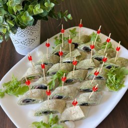 Szama-nka Catering - Elegancko podane wrapy z oliwkami i rukolą na białym talerzu, ozdobione czerwonymi wykałaczkami, obok dekoracyjny kwiat w doniczce. Idealne na catering firmowy.