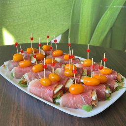 Szama-nka Catering - Eleganckie roladki z szynki parmeńskiej, brie i pomidorków koktajlowych na białym talerzu, dekorowane wykałaczkami z czerwonymi kostkami. Tło: zielone liście.
