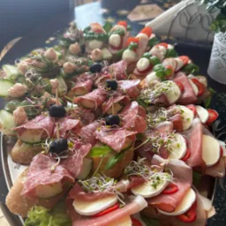 Elegancka taca z różnorodnymi kanapkami: salami, mozzarella z pomidorem i bazylią, ogórek z pastą, udekorowane kiełkami. Idealne na catering firmowy.
