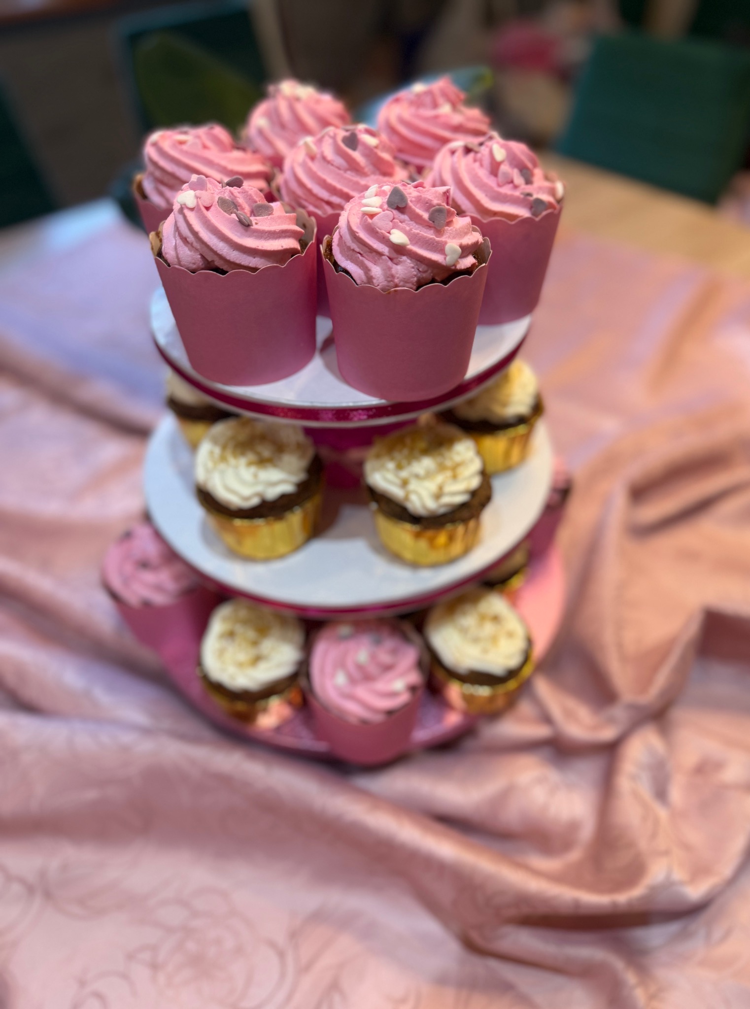 Dwupoziomowa patera z babeczkami: różowe z serduszkami na górze, złote z białym kremem na dole, całość na różowym obrusie. Słodki stół.