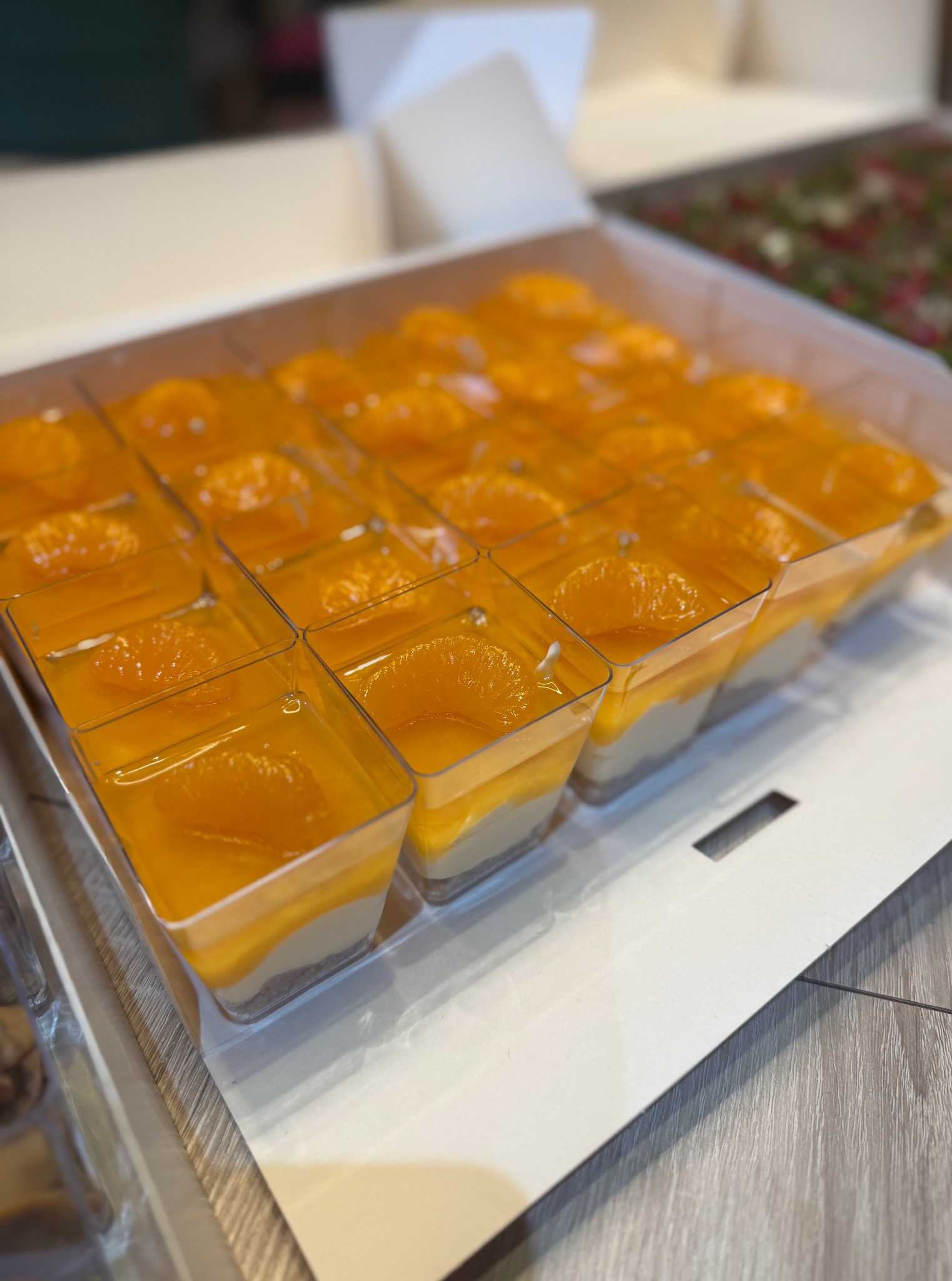 Deser w przezroczystych pojemnikach, ułożony w kartonowym pudełku. Widoczne warstwy: biszkopt, krem i galaretka z kawałkiem pomarańczy.