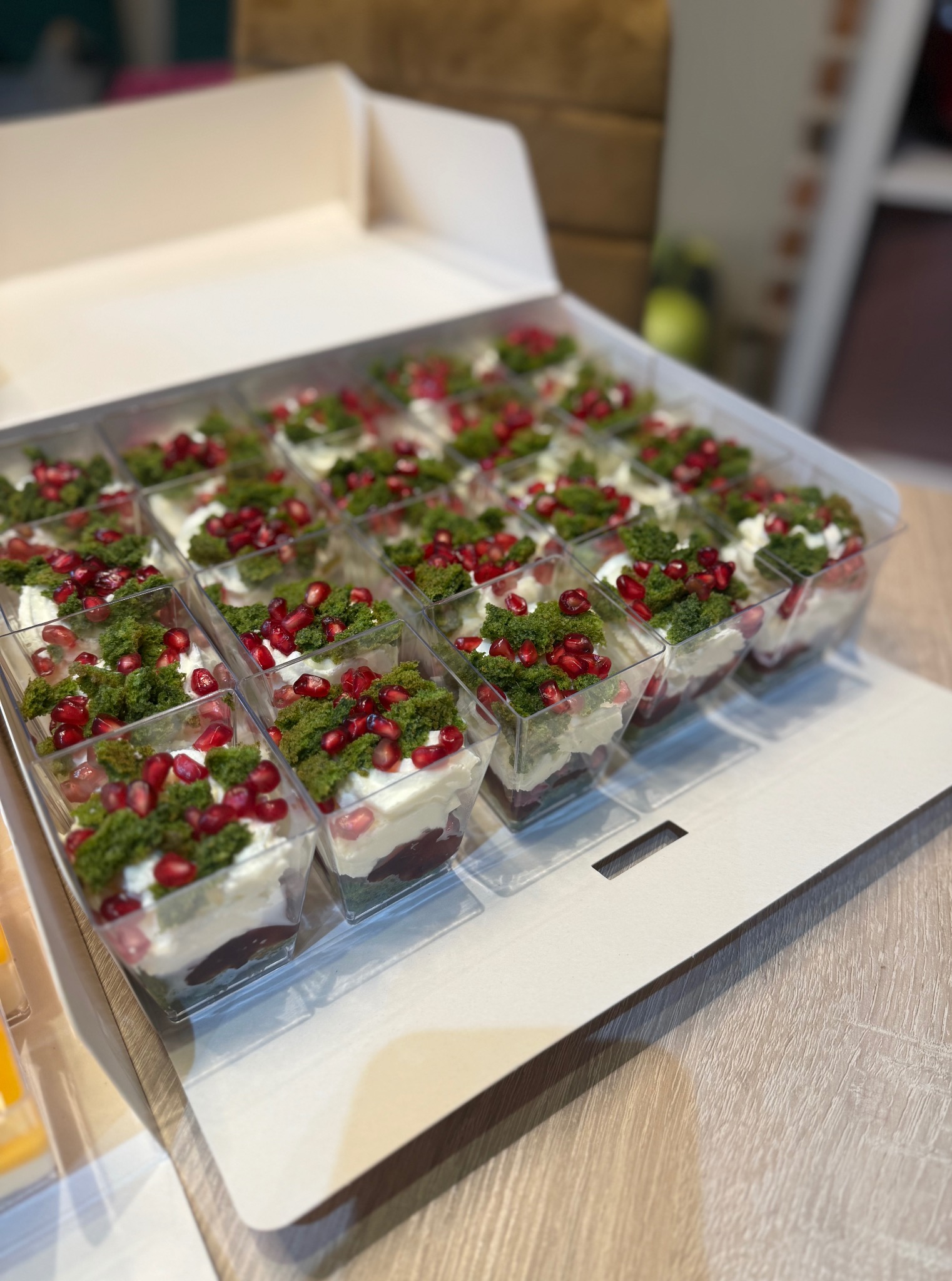 Eleganckie, świąteczne desery w przezroczystych pojemnikach, ułożone w białym pudełku. Desery udekorowane kremem, zielonym biszkoptem i granatem.