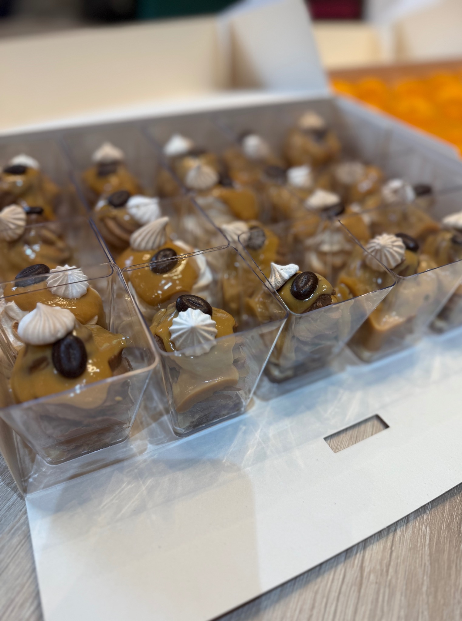 Kremowe mini desery w przezroczystych pojemnikach, udekorowane bezą i ziarnem kawy, ułożone w białym pudełku. Eleganckie słodkości na catering.