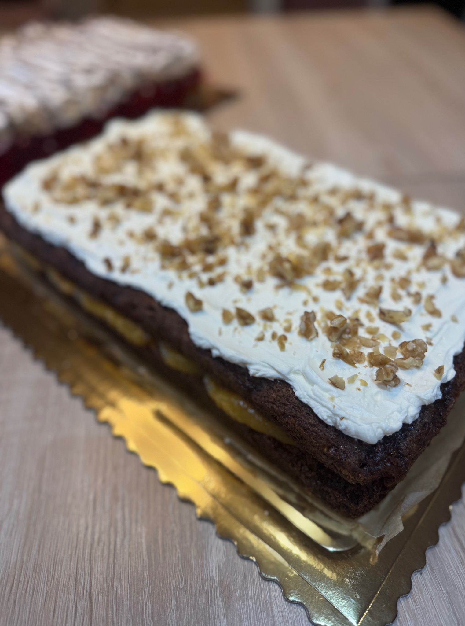Prostokątny tort z kremem i orzechami na złotym podkładzie. Widoczna struktura ciasta i kremu. W tle inny tort.