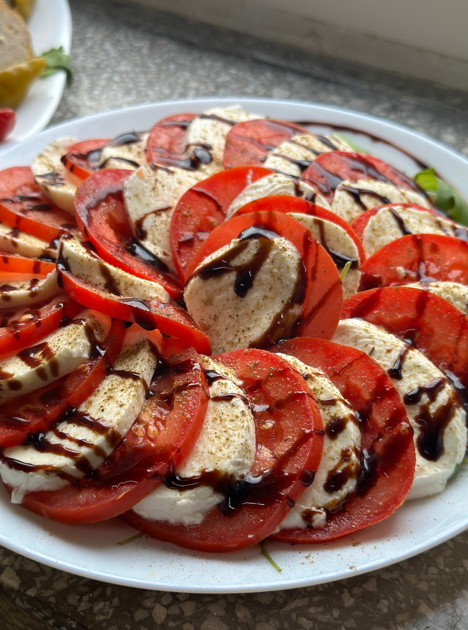 Apetyczna sałatka Caprese na białym talerzu, skropiona octem balsamicznym. Pomidory i mozzarella ułożone naprzemiennie. Delikatne przyprawy posypane na wierzchu.