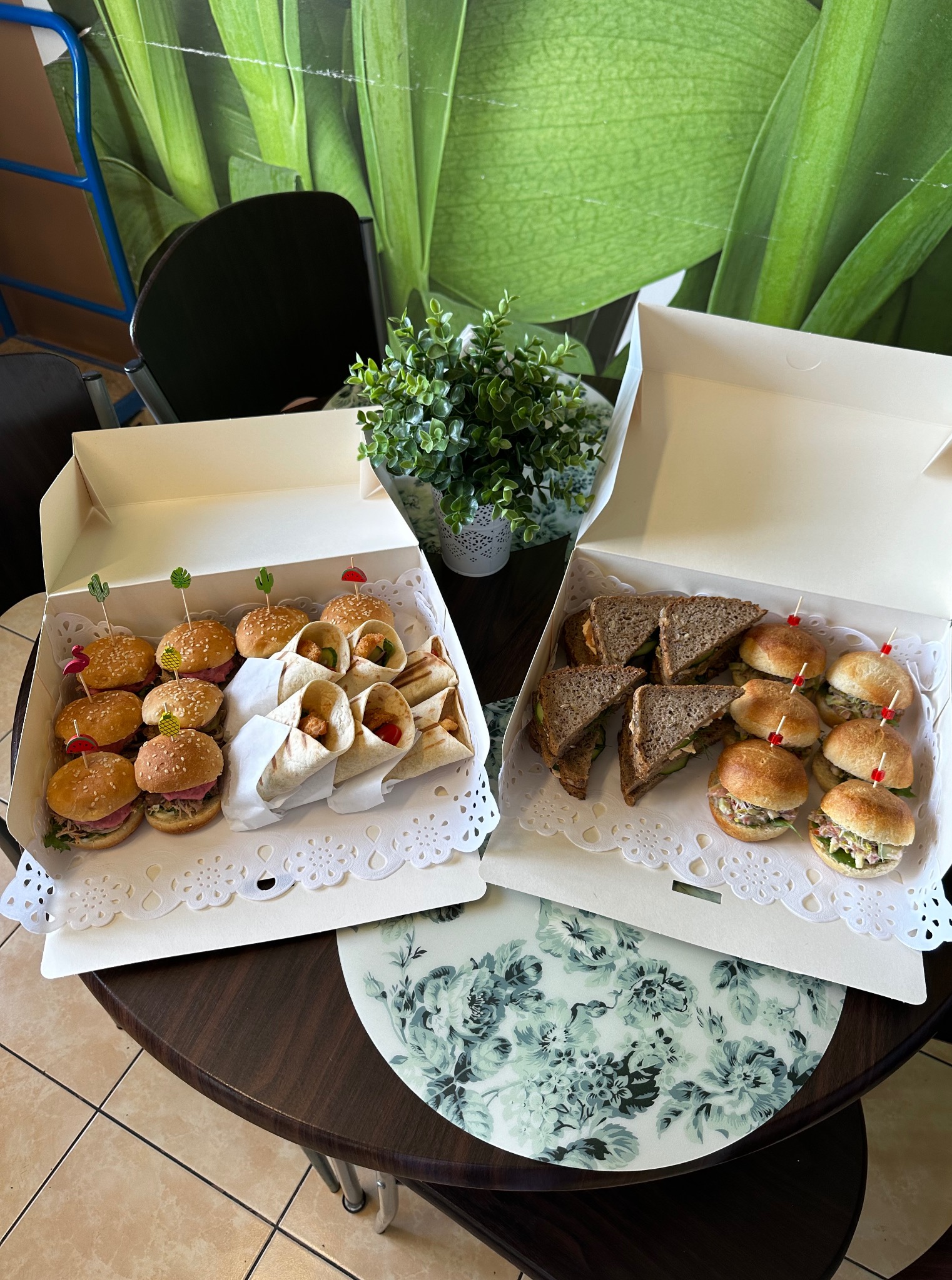 Dwie białe kartonowe tace z mini burgerami, wrapami i kanapkami na okrągłym stole z kwiatowym wzorem, tło z zielonym motywem roślinnym.