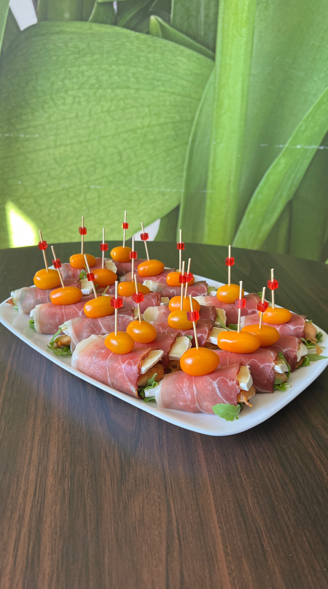 Eleganckie roladki z szynki parmeńskiej, brie i pomidorków koktajlowych na białym talerzu, dekorowane wykałaczkami z czerwonymi kostkami. Tło: zielone liście.