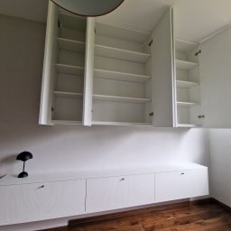 MODERNO Agnieszka Wieczorek - Biała szafa wnękowa z otwartymi górnymi szafkami, minimalistyczny design, drewniana podłoga, czarna lampka na szafce. Widoczne zawiasy i półki.