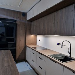 MODERNO Agnieszka Wieczorek - Nowoczesna kuchnia z drewnianymi i białymi szafkami, czarnym zlewem i płytą indukcyjną. Uwagę przyciąga niedokończony sufit i zabezpieczona podłoga.