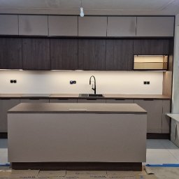 MODERNO Agnieszka Wieczorek - Nowoczesna kuchnia z wyspą w odcieniach beżu i brązu, oświetlenie LED pod szafkami, widok na okno i drzwi balkonowe, zabezpieczona folią ochronną podłoga.