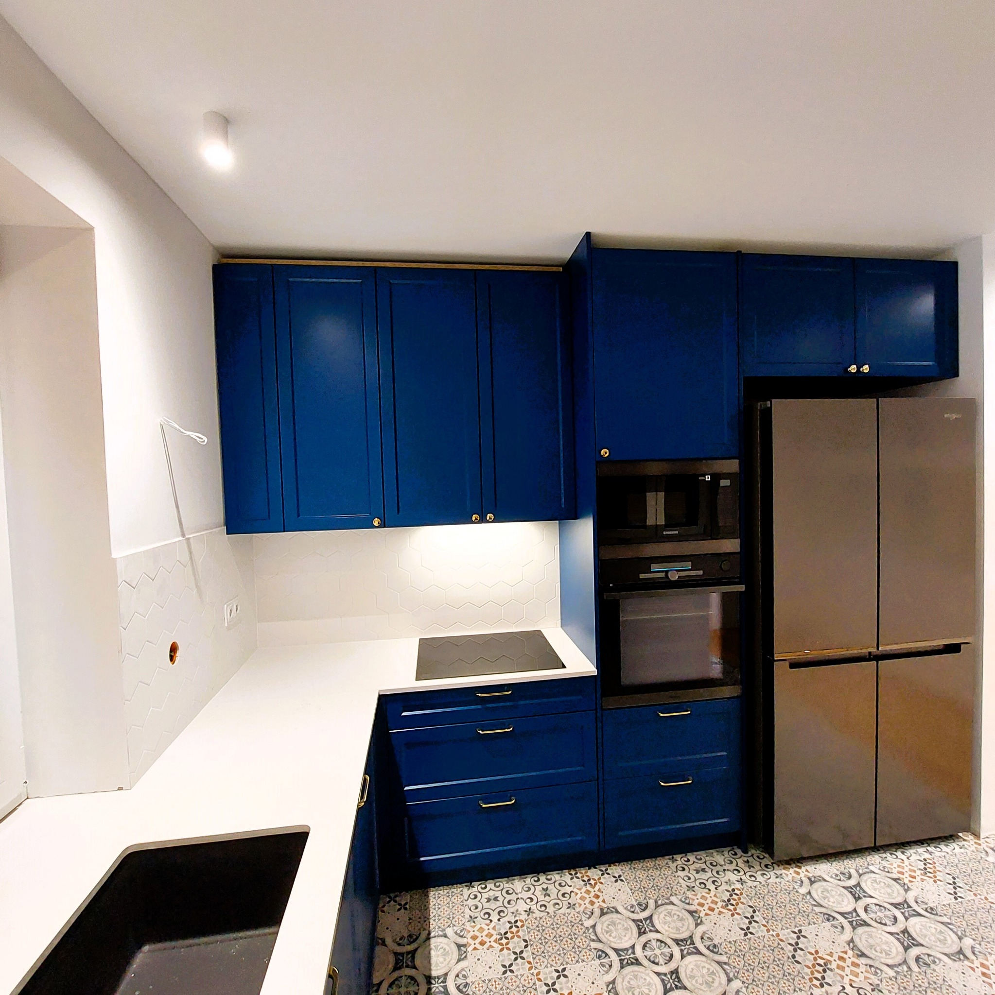 Niebieska zabudowa kuchenna z frezowanymi frontami, złote uchwyty, biały blat i heksagonalne płytki nad blatem. Na podłodze wzorzysta mozaika.