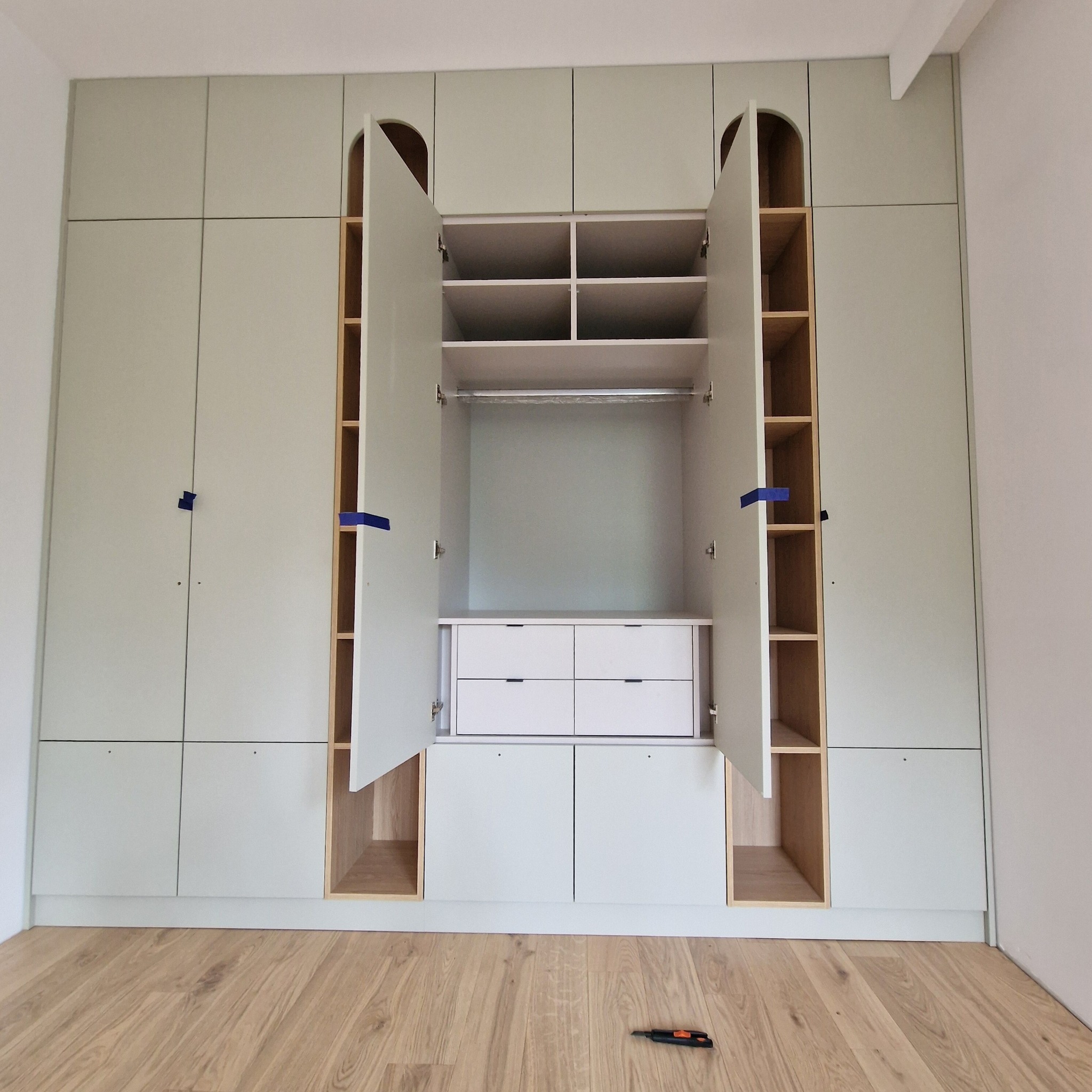 Jasnozielona szafa wnękowa z otwartymi drzwiami, ukazująca półki i szuflady. Na podłodze leży nóż introligatorski.