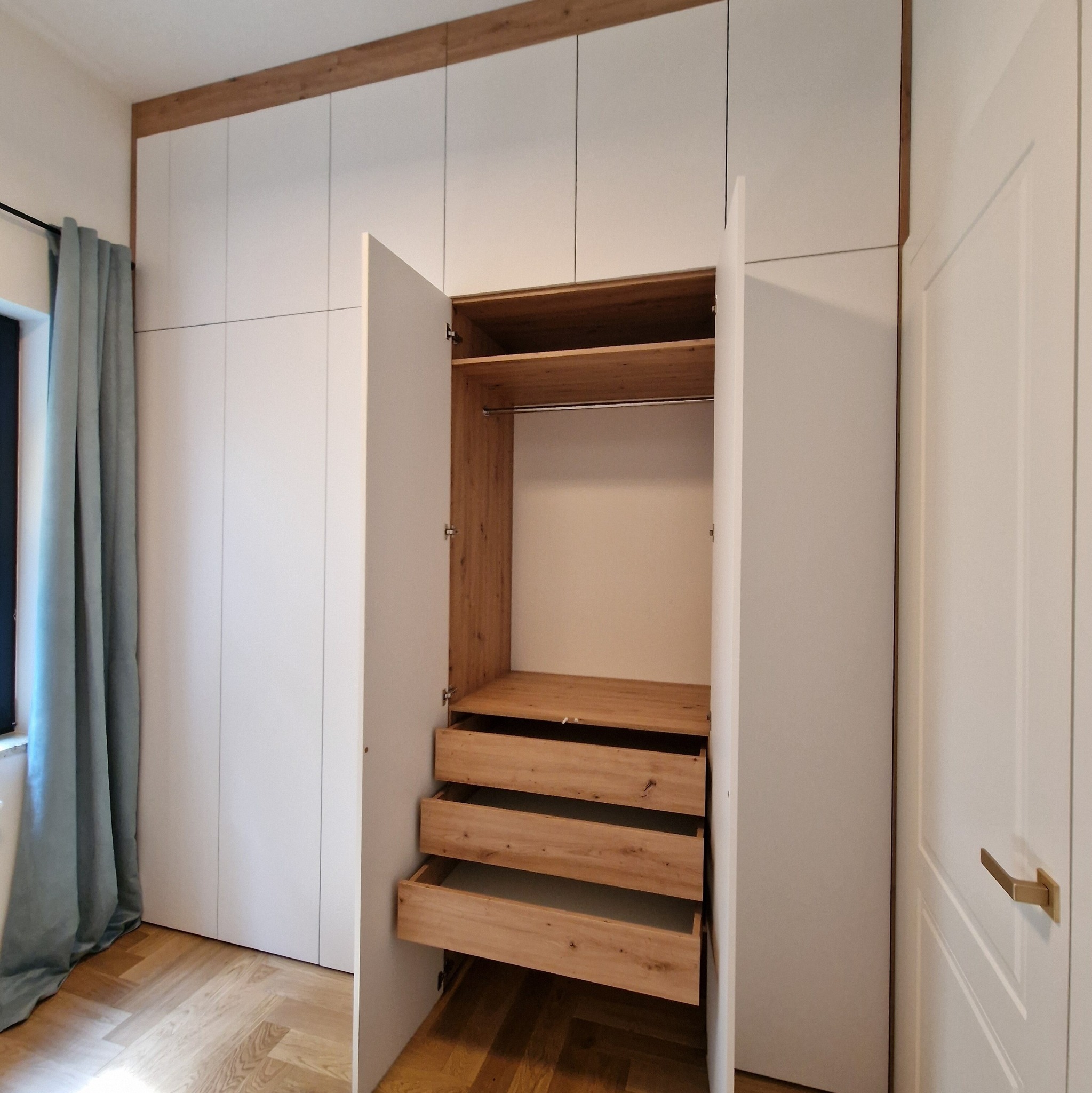 Biała szafa wnękowa z drewnianymi elementami, częściowo otwarta, ukazująca szuflady i półki. Widoczny parkiet i zasłona w tle.