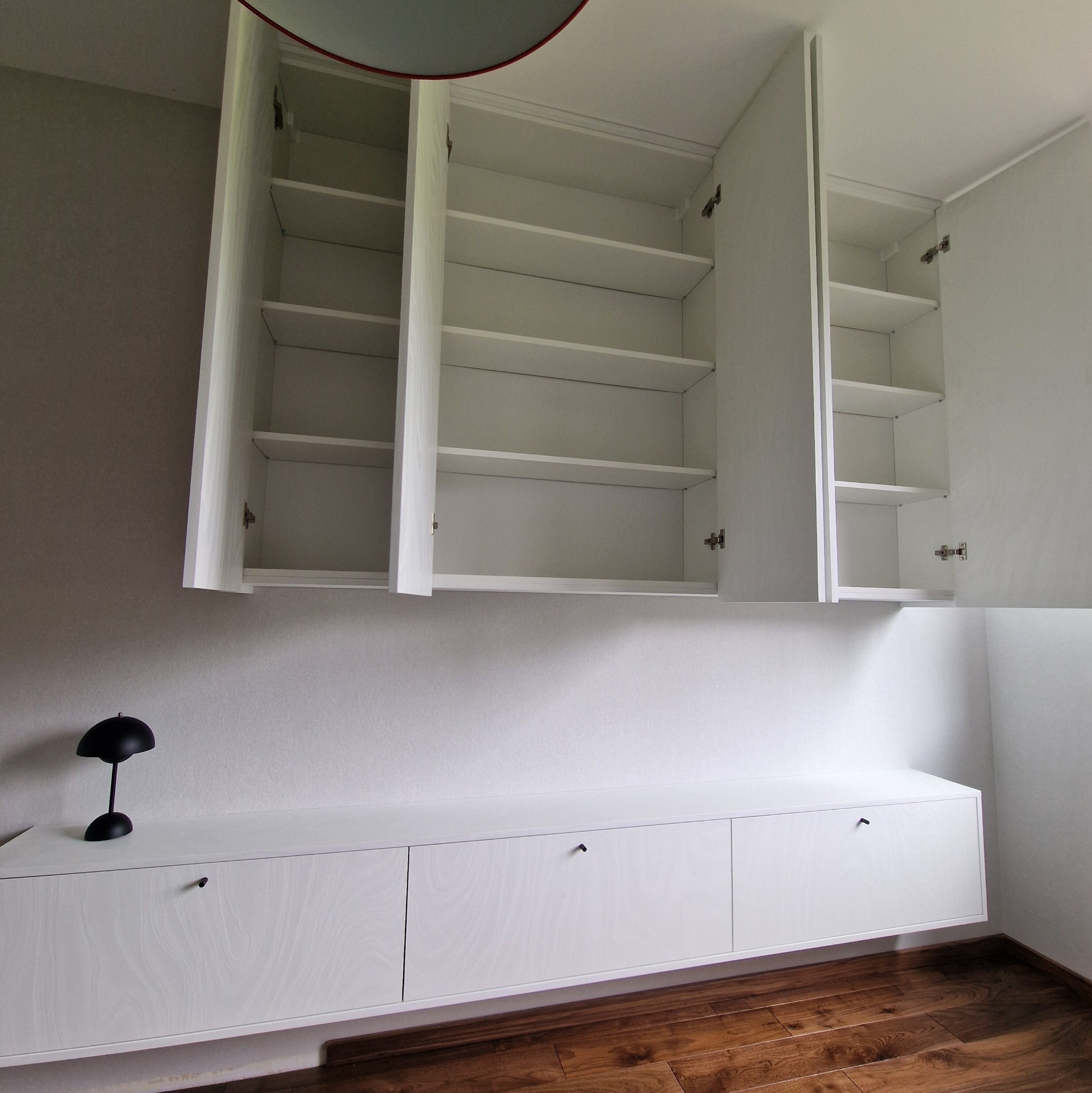 Biała szafa wnękowa z otwartymi górnymi szafkami, minimalistyczny design, drewniana podłoga, czarna lampka na szafce. Widoczne zawiasy i półki.