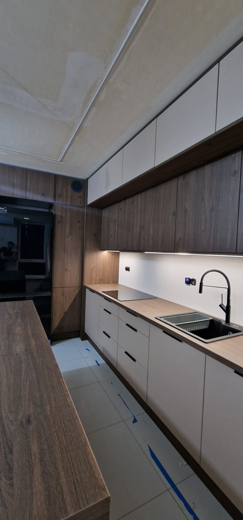 Nowoczesna kuchnia z drewnianymi i białymi szafkami, czarnym zlewem i płytą indukcyjną. Uwagę przyciąga niedokończony sufit i zabezpieczona podłoga.