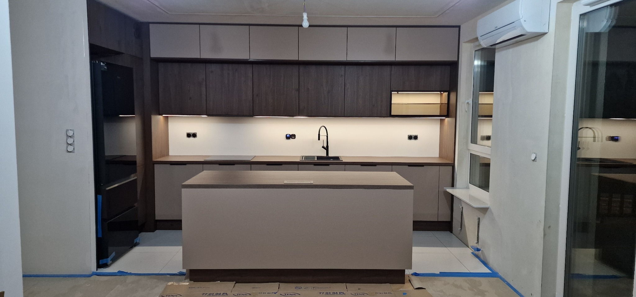 Nowoczesna kuchnia z wyspą w odcieniach beżu i brązu, oświetlenie LED pod szafkami, widok na okno i drzwi balkonowe, zabezpieczona folią ochronną podłoga.