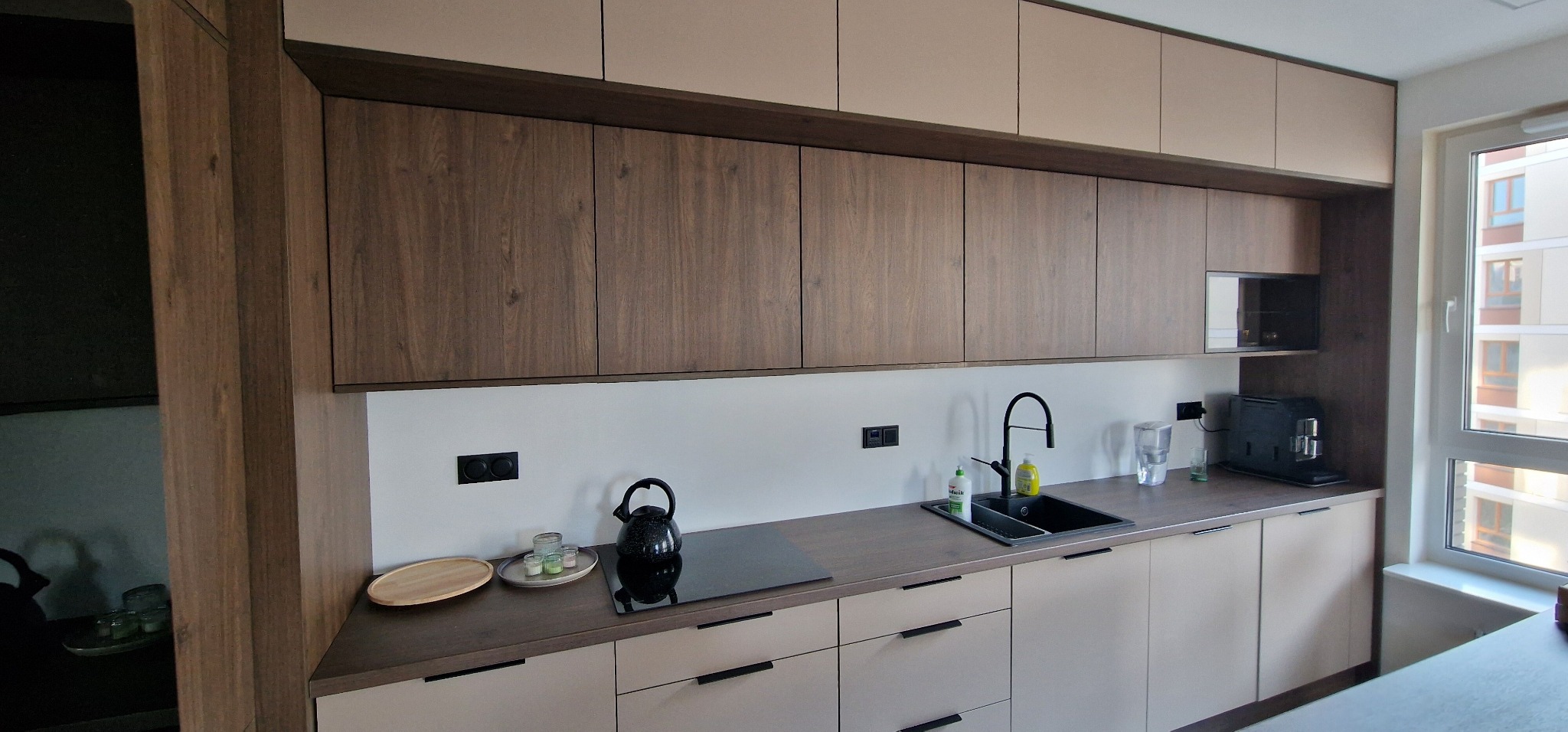 Nowoczesna kuchnia z drewnianymi i beżowymi frontami, czarnymi detalami i sprzętami. Widok na okno z budynkiem w tle. Minimalistyczny design.