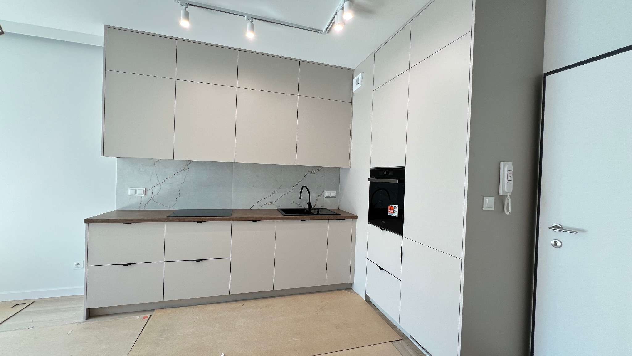 Jasna kuchnia w kształcie litery L z frontami z płyty kaszmirowej, drewnopodobnym blatem i czarną armaturą. Widoczna zabudowa piekarnika i płyta indukcyjna.