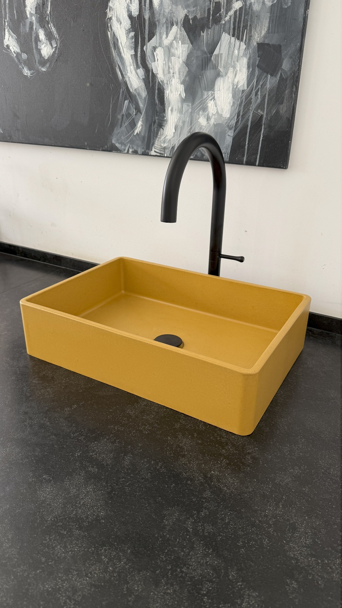 Żółta, prostokątna umywalka na czarnym blacie pod abstrakcyjnym obrazem. Nowoczesna armatura w matowej czerni. Minimalistyczny design łazienki.