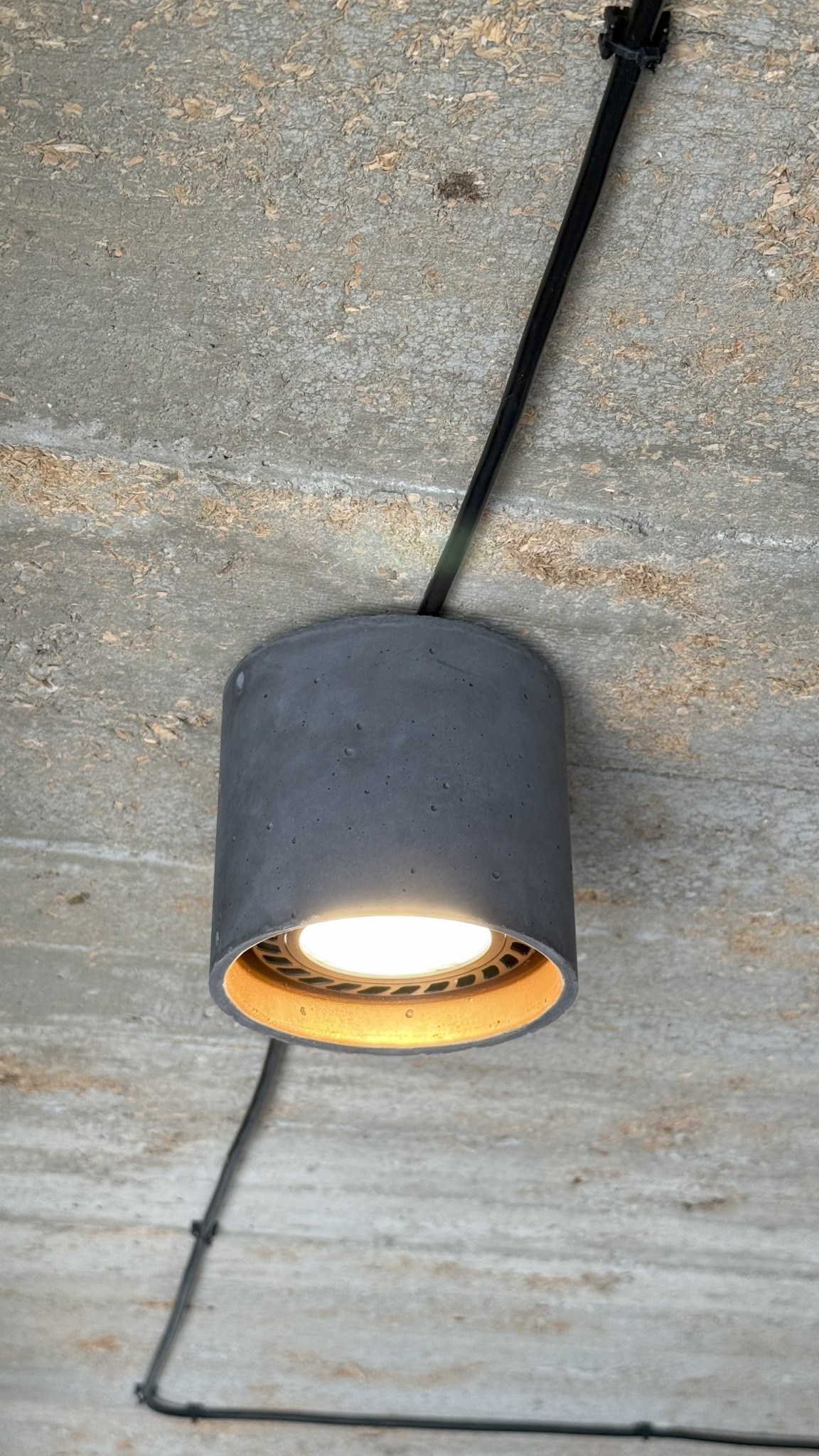 Betonowa lampa sufitowa z ciepłym światłem, czarny kabel biegnie wzdłuż surowego betonowego sufitu z widoczną fakturą i naturalnymi przebarwieniami.