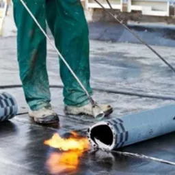 Pracownik w zielonych spodniach spawa papę termozgrzewalną na dachu płaskim. Widoczne rolki papy i palnik z płomieniem. Praca dekarska w toku.