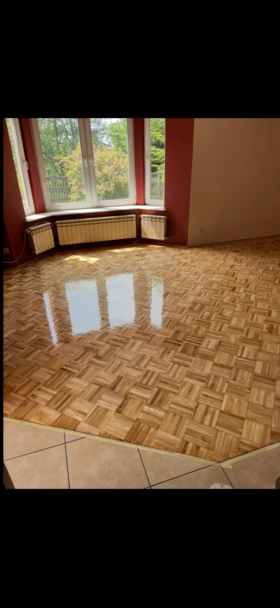 Parkiet po renowacji w pokoju z oknem i grzejnikami. Podłoga błyszczy, odbijając światło. Widoczne zabezpieczenia taśmą malarską przy łączeniu z płytkami.