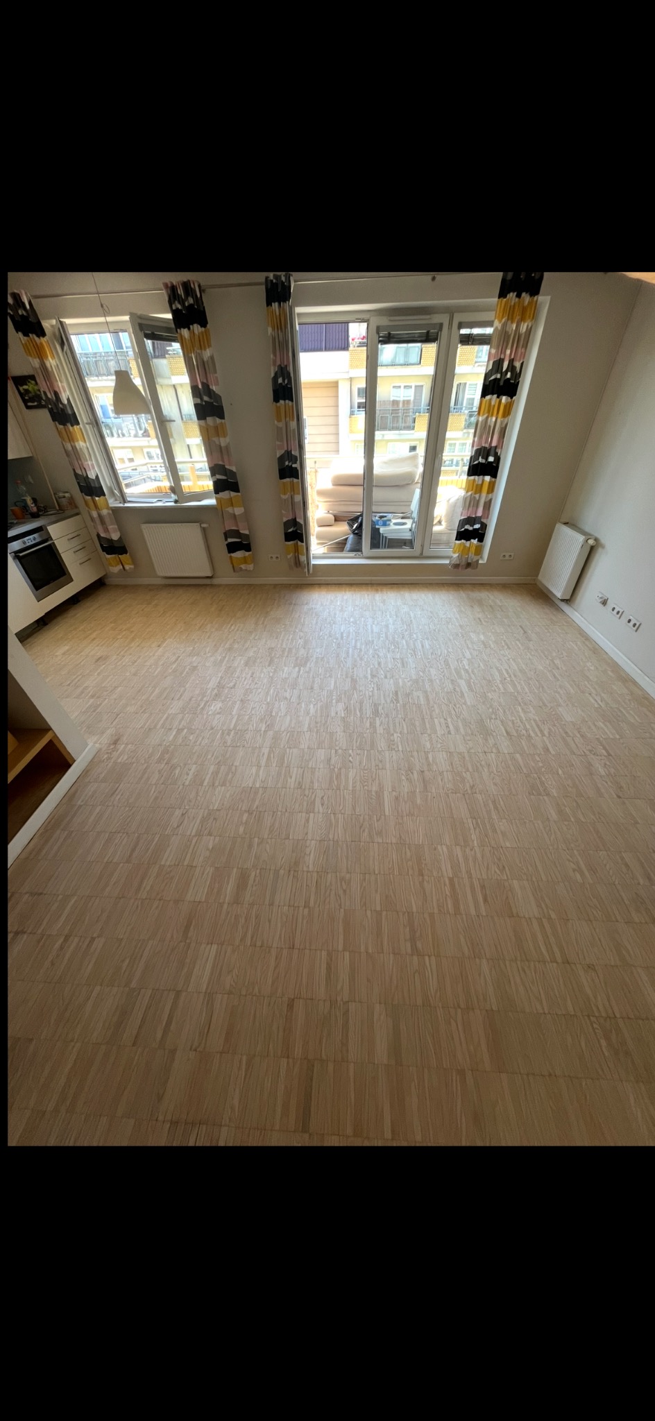 Jasna, odnowiona podłoga parkietowa w przestronnym pokoju z oknami balkonowymi i wzorzystymi zasłonami. Widoczny fragment kuchni i grzejnik.