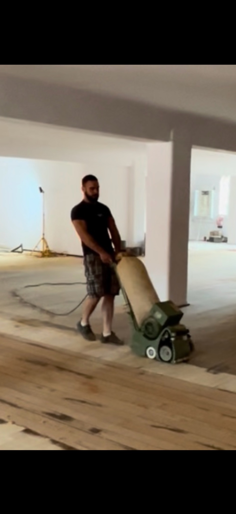 Mężczyzna obsługuje maszynę do renowacji podłogi, widoczne świeżo odnowione deski w jasnym pomieszczeniu z kolumnami. Praca w toku.