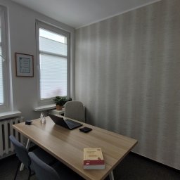 MG Finanse Monika Górna - Jasne biuro z biurkiem, laptopem, kalkulatorem i kodeksem. Na ścianie certyfikat. Dwa okna z żaluzjami. Minimalistyczny wystrój wnętrza.