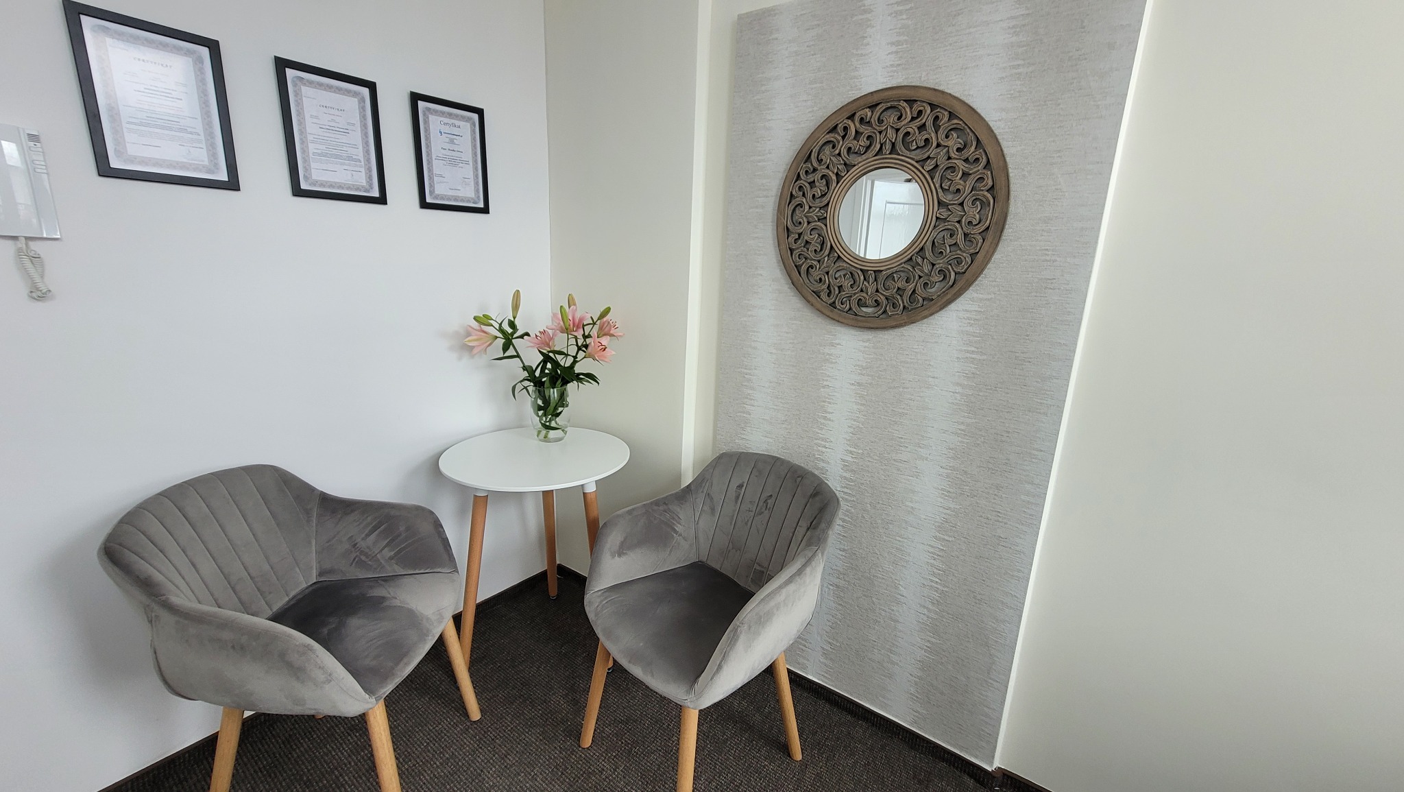 Wnętrze biura rachunkowego w Gdyni: dwa szare krzesła dla klientów, stolik z kwiatami, certyfikaty na ścianie i okrągłe lustro dekoracyjne.