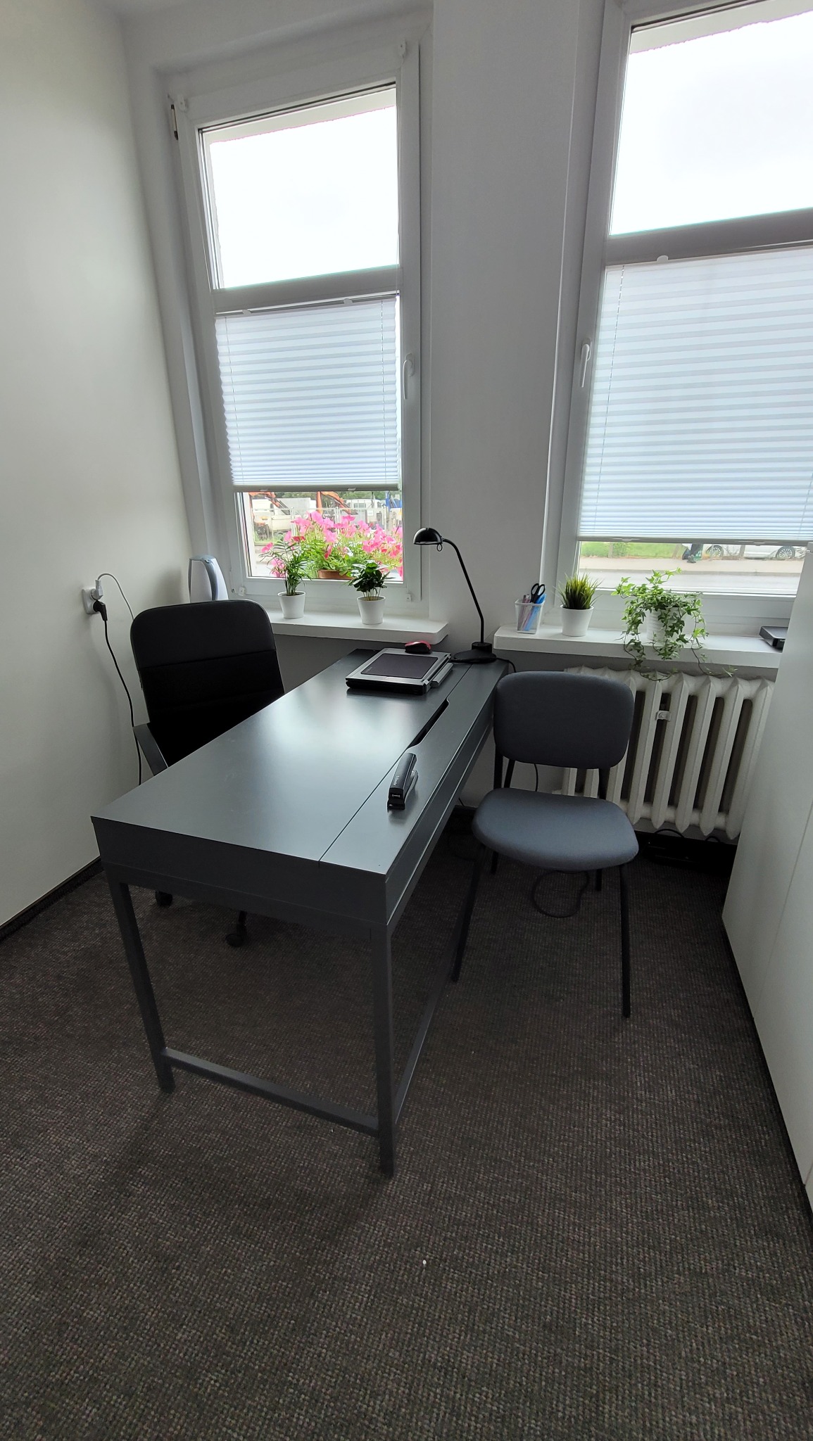 Nowoczesne biuro: szare biurko z lampką i tabletem na tle okien z żaluzjami, kwiaty w doniczkach. Minimalistyczny design, stonowana kolorystyka, przytulne miejsce pracy.