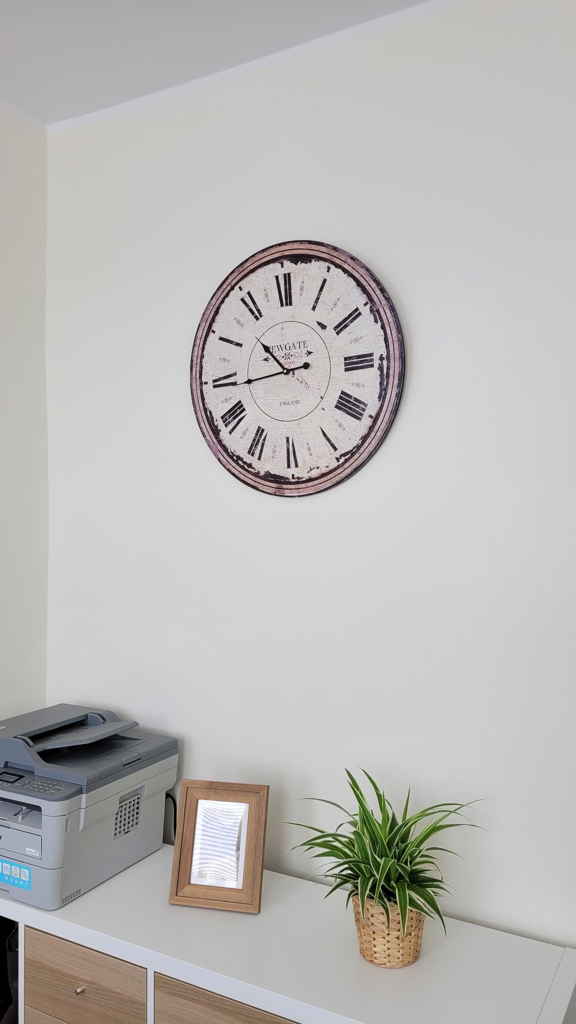 Biuro: zegar na ścianie nad biurkiem z drukarką, ramką na zdjęcia i rośliną doniczkową. Jasne wnętrze, neutralne kolory, spokojna atmosfera pracy.
