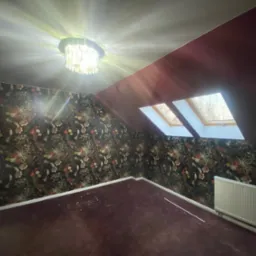 Pokój na poddaszu z oknami dachowymi i ozdobną tapetą w kwiaty. Centralnie umieszczony żyrandol oświetla wnętrze.