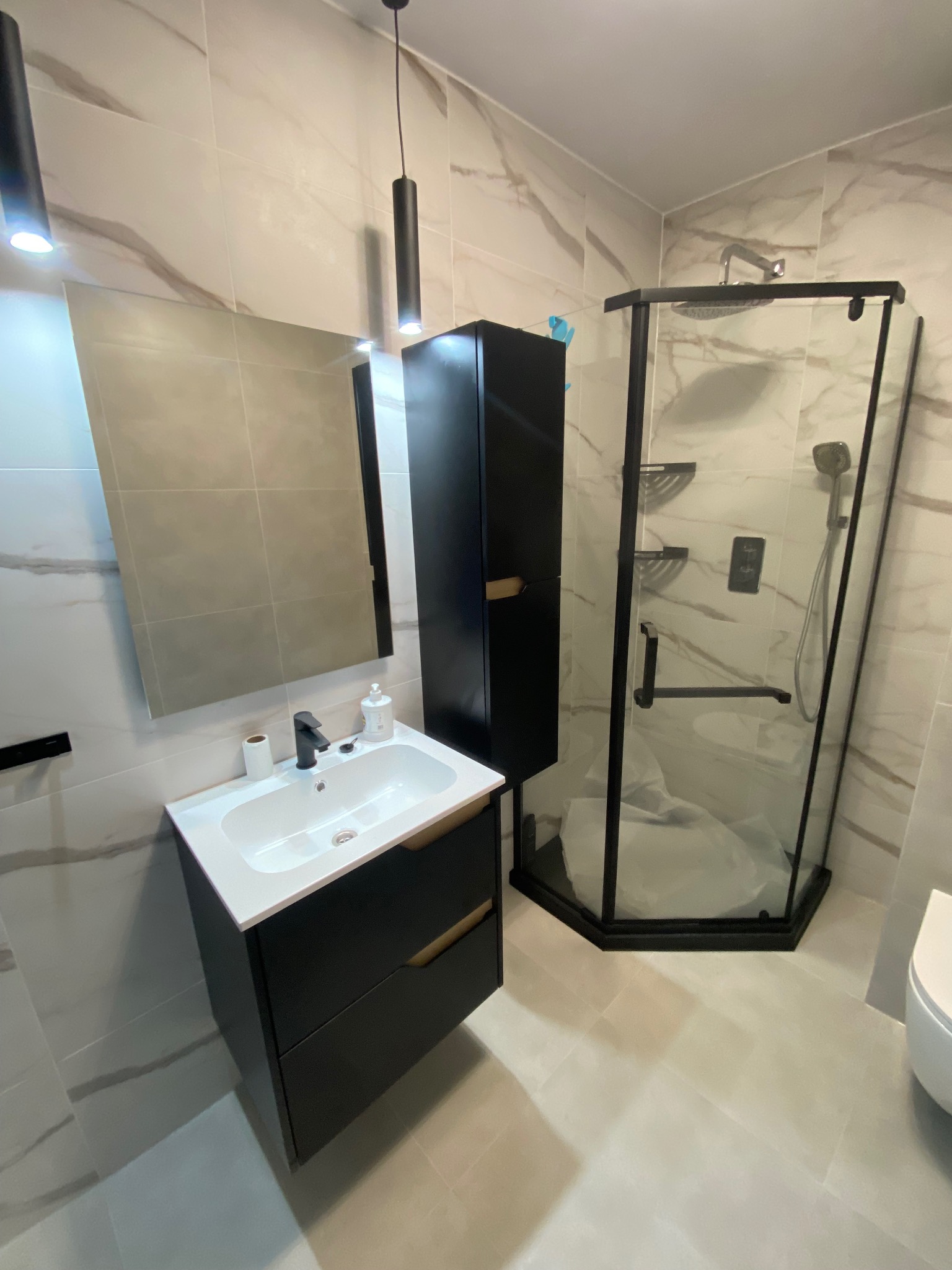 Nowoczesna łazienka po remoncie: kabina prysznicowa w czarnej ramie, umywalka z szafką w kolorze czarnym, marmurowe płytki, minimalistyczne oświetlenie.