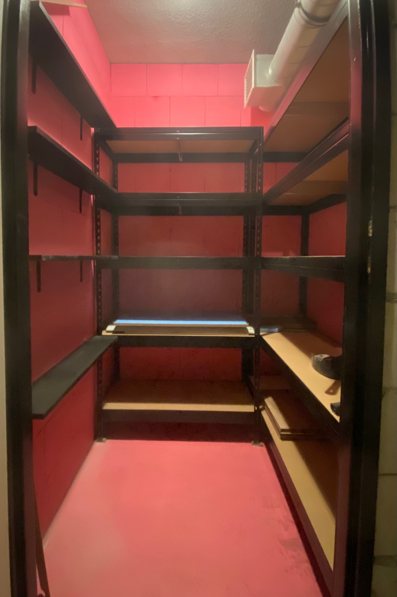 Różowe pomieszczenie gospodarcze z metalowymi regałami o różnej konfiguracji, oświetlone górnym światłem.