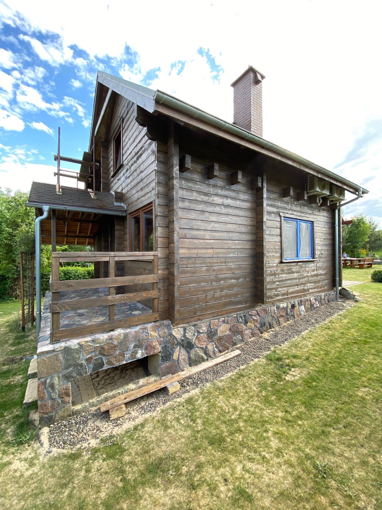 Drewniany dom z kamienną podmurówką i zadaszonym tarasem w trakcie prac wykończeniowych, widoczne rusztowania i zabezpieczone okno folią malarską.