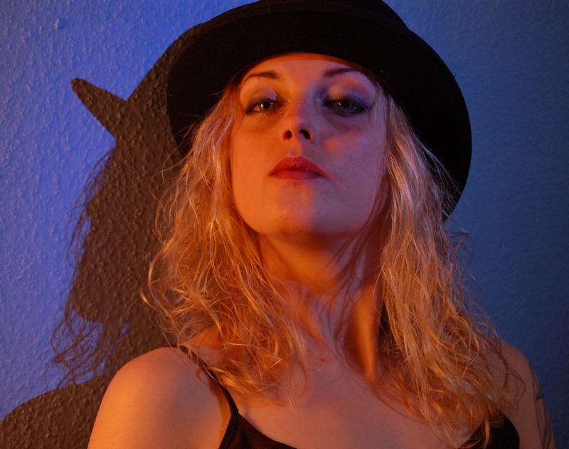 Portret kobiety w kapeluszu na tle niebieskiej ściany z cieniem. Ujęcie z dołu, ciepłe oświetlenie, podkreślone rysy twarzy.