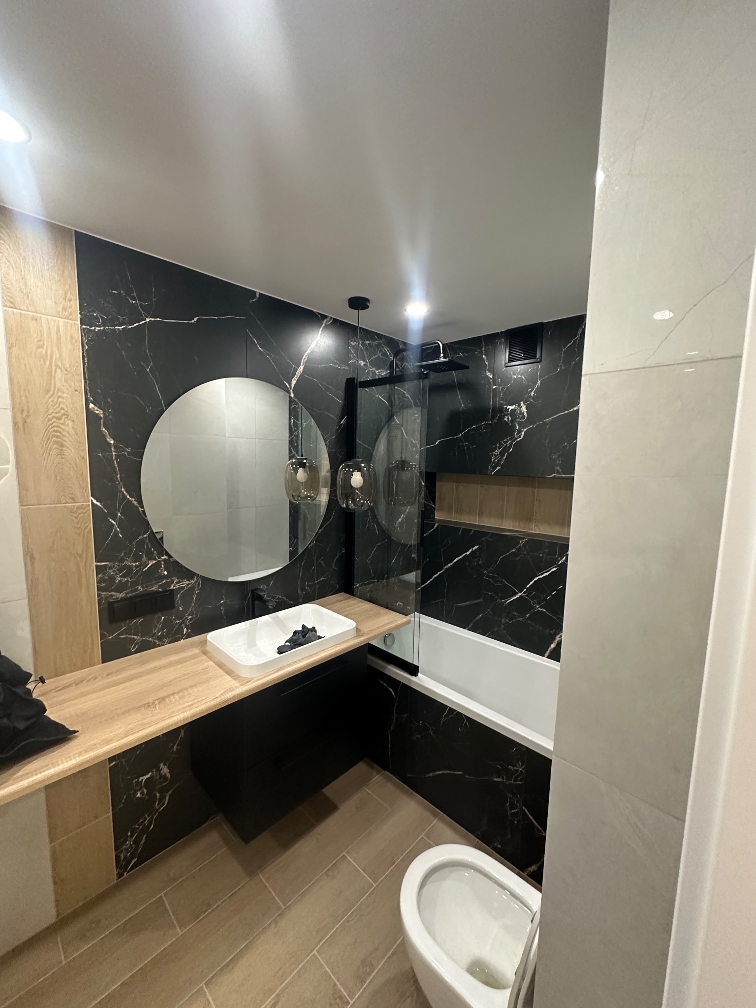 Nowoczesna łazienka z czarnymi płytkami imitującymi marmur, okrągłym lustrem i drewnianym blatem pod umywalką. Widoczna wanna i toaleta.