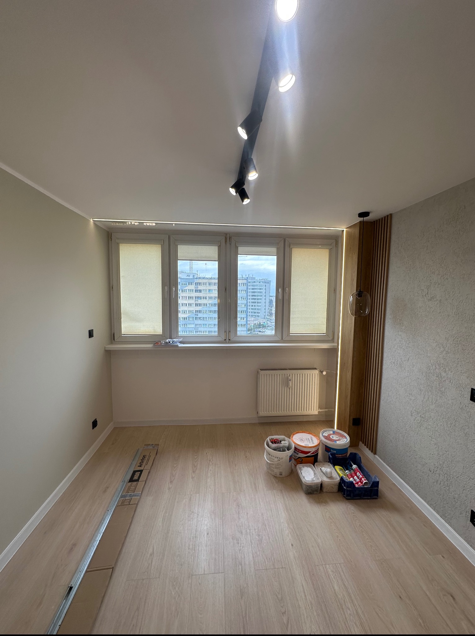 Jasne wnętrze pokoju po remoncie: świeżo pomalowane ściany, zamontowane rolety w oknach, nowe panele podłogowe i nowoczesne oświetlenie sufitowe.