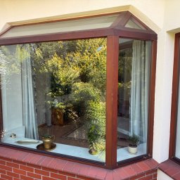 Volodymyr Storozum ViOS - Kątowe okno z brązowymi ramami, widok na zieleń z zewnątrz, parapet z cegły i rośliny doniczkowe. Widoczne firanki w oknach.