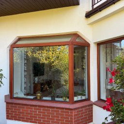 Volodymyr Storozum ViOS - Wystający, przeszklony wykusz okienny z drewnianą ramą, osadzony na ceglanym murku, z widokiem na wnętrze i zieleń za oknem, oraz rosnące czerwone kwiaty po prawej stronie.