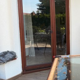 Volodymyr Storozum ViOS - Brązowe dwuskrzydłowe drzwi balkonowe w trakcie renowacji. Widoczna rama okienna na stojaku, narzędzie i worek na odpady. Tło: biała ściana i zieleń.