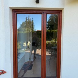 Volodymyr Storozum ViOS - Brązowe, dwuskrzydłowe okno balkonowe z widocznym odbiciem ogrodu i osoby, osadzone w białej elewacji z lampą. Widoczna podłoga z płytek imitujących drewno.