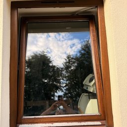 Volodymyr Storozum ViOS - Drewniane okno z widocznym uszkodzeniem ramy i śladami farby. W szybie odbija się niebo i drzewa, a także osoba robiąca zdjęcie telefonem.