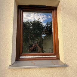 Volodymyr Storozum ViOS - Drewniane okno z widocznym odbiciem osoby robiącej zdjęcie na tle błękitnego nieba i zieleni. Detal elewacji z jasnym, chropowatym tynkiem.