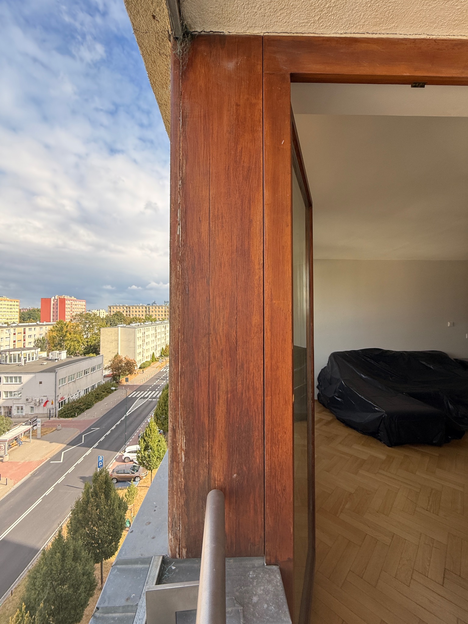 Otwarta, drewniana rama okienna z widokiem na miasto z balkonu. Wnętrze pokoju z parkietem zabezpieczone folią malarską.